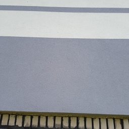 Złota rączka - Fragment elewacji budynku z szarą i białą farbą w poziome pasy oraz ozdobnym elementem dekoracyjnym przy podstawie ściany.