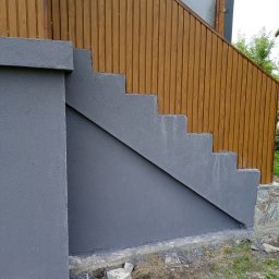 Złota rączka - Zewnętrzne betonowe schody pomalowane na szaro, przylegające do elewacji z paneli drewnopodobnych. Widoczne niedociągnięcia przy podstawie schodów i resztki materiałów budowlanych.