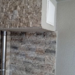 Złota rączka - Ściana wykończona dekoracyjnym kamieniem w odcieniach szarości, z widocznym kominem i białą klapą rewizyjną w rogu pomieszczenia.
