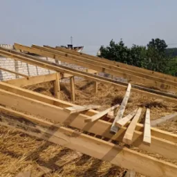 Konstrukcja więźby dachowej z drewnianych belek na poddaszu, widoczne fragmenty muru z białej cegły i słoma, niebieskie niebo w tle.