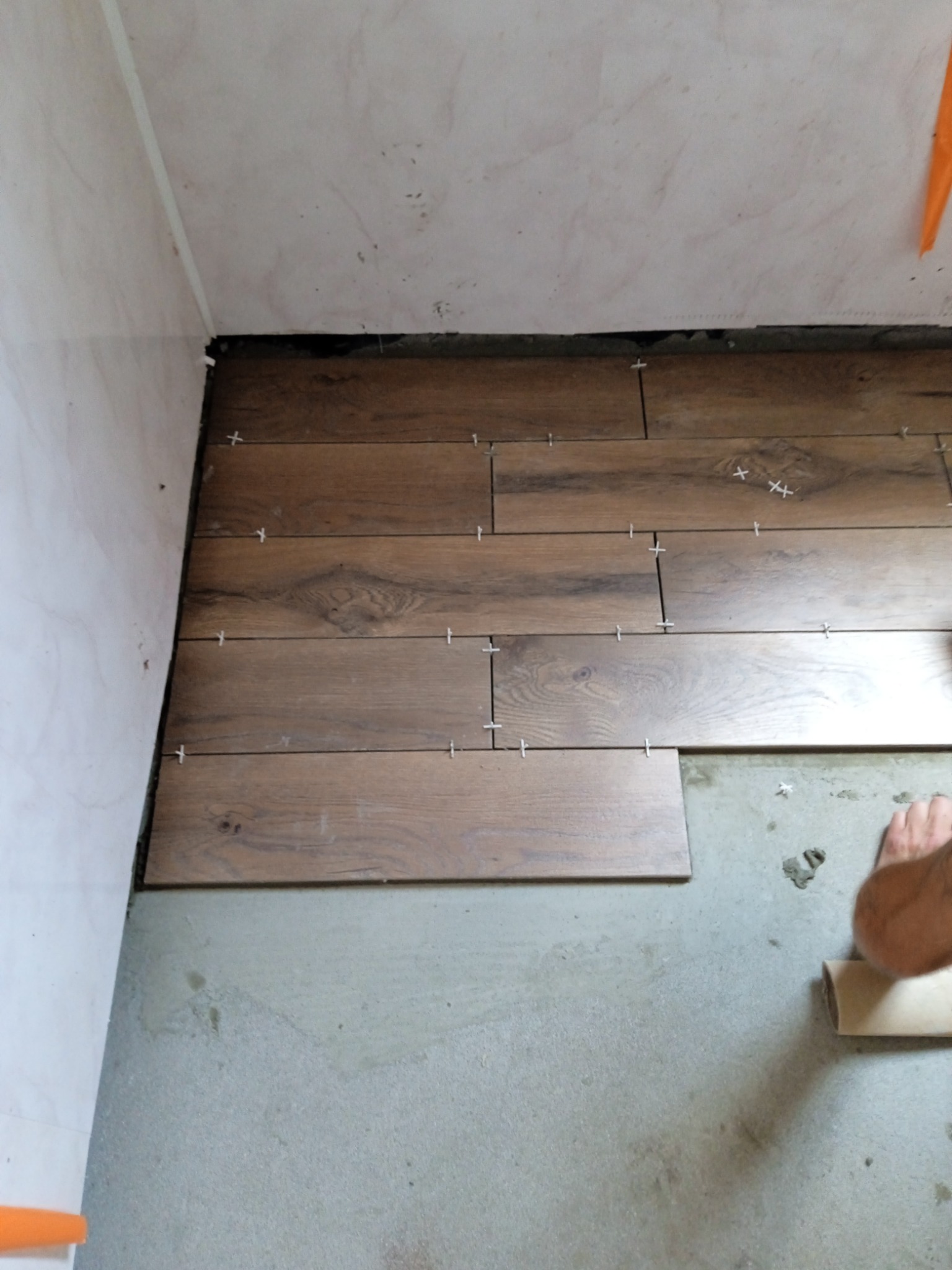Układanie drewnopodobnych płytek podłogowych z użyciem krzyżyków dystansowych; widoczny fragment stopy i rolka taśmy malarskiej.