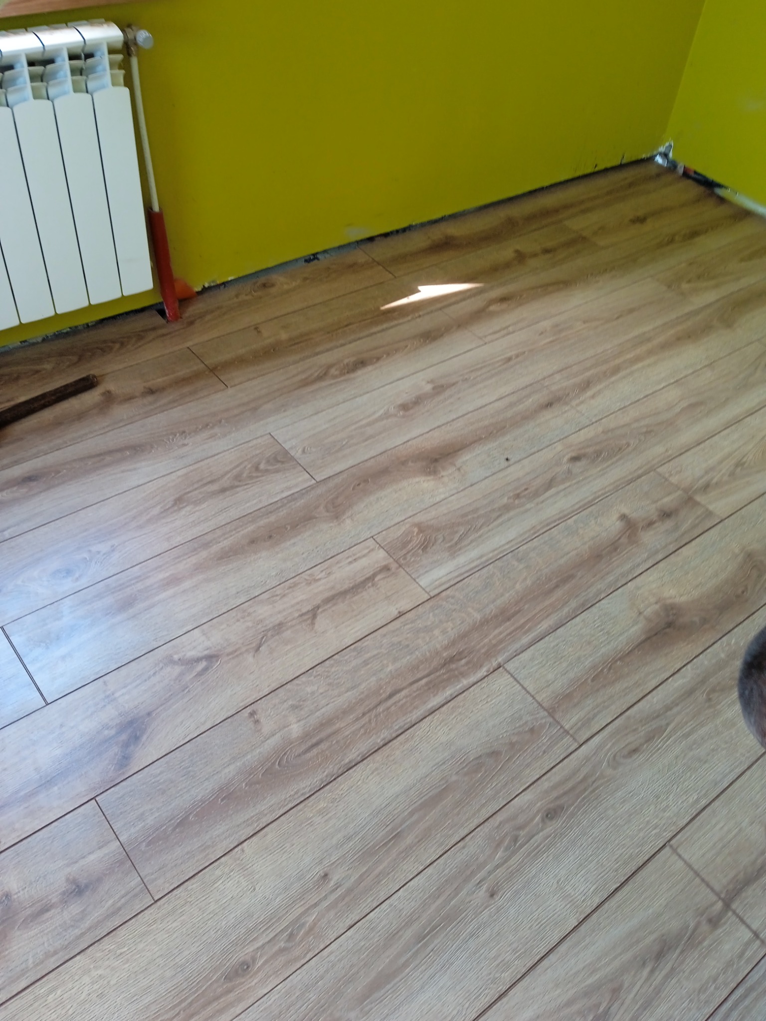 Ułożona podłoga z paneli laminowanych w jasnym odcieniu drewna, widoczny grzejnik i żółta ściana w tle.