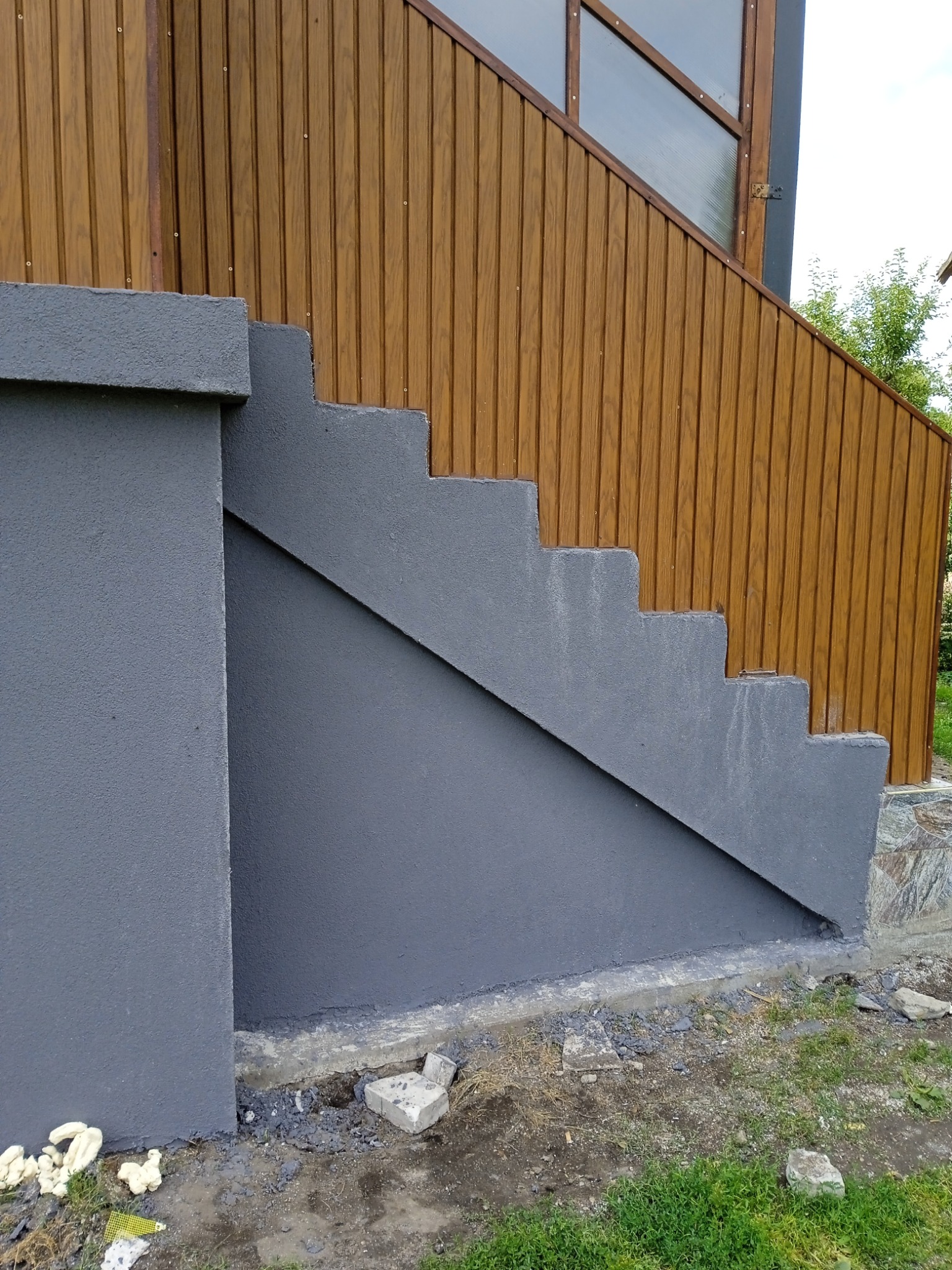 Zewnętrzne betonowe schody pomalowane na szaro, przylegające do elewacji z paneli drewnopodobnych. Widoczne niedociągnięcia przy podstawie schodów i resztki materiałów budowlanych.