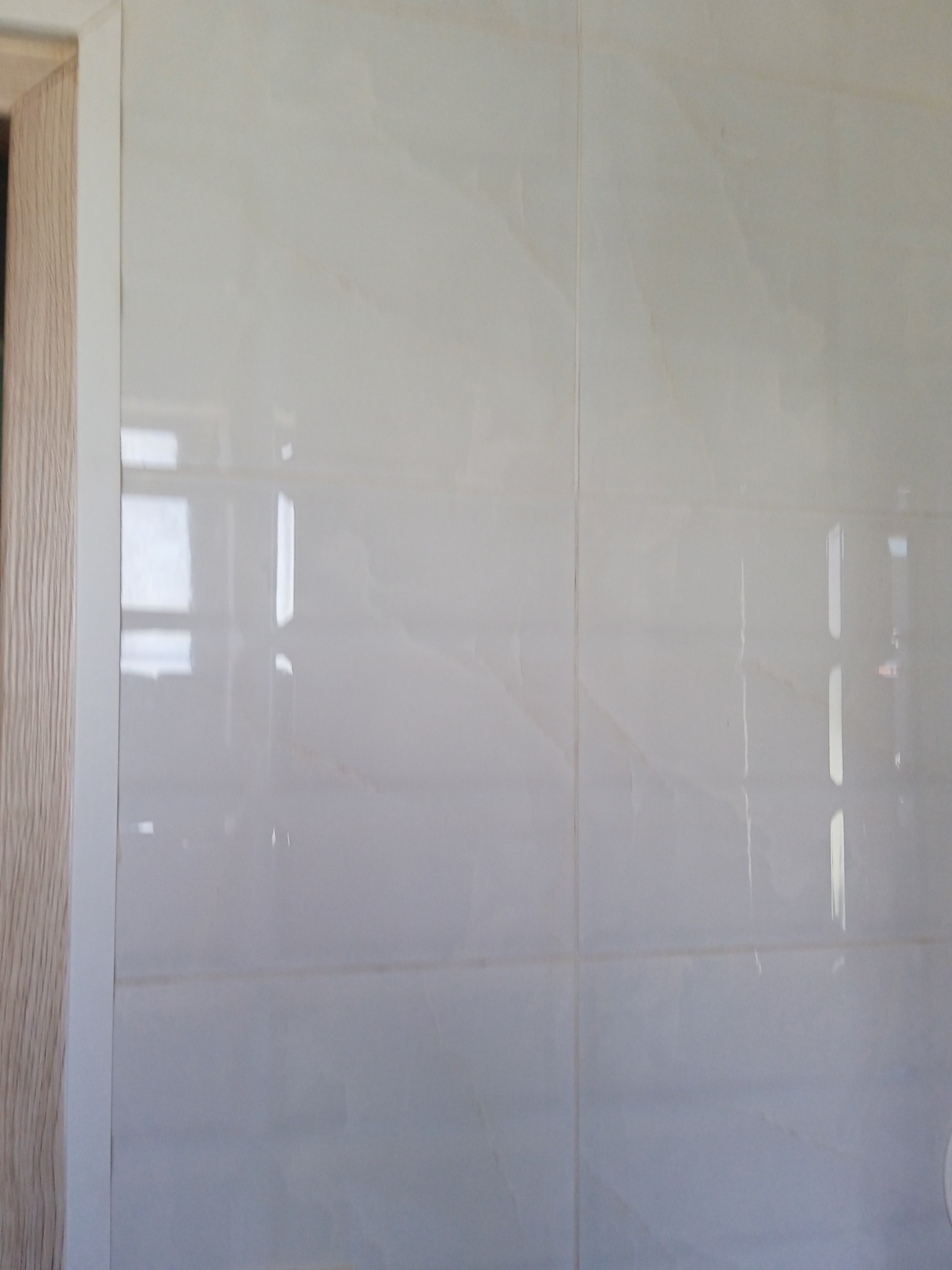 Ściana wyłożona jasnymi, błyszczącymi płytkami imitującymi marmur w Mężeninie, widoczny fragment drewnianej ościeżnicy drzwi.