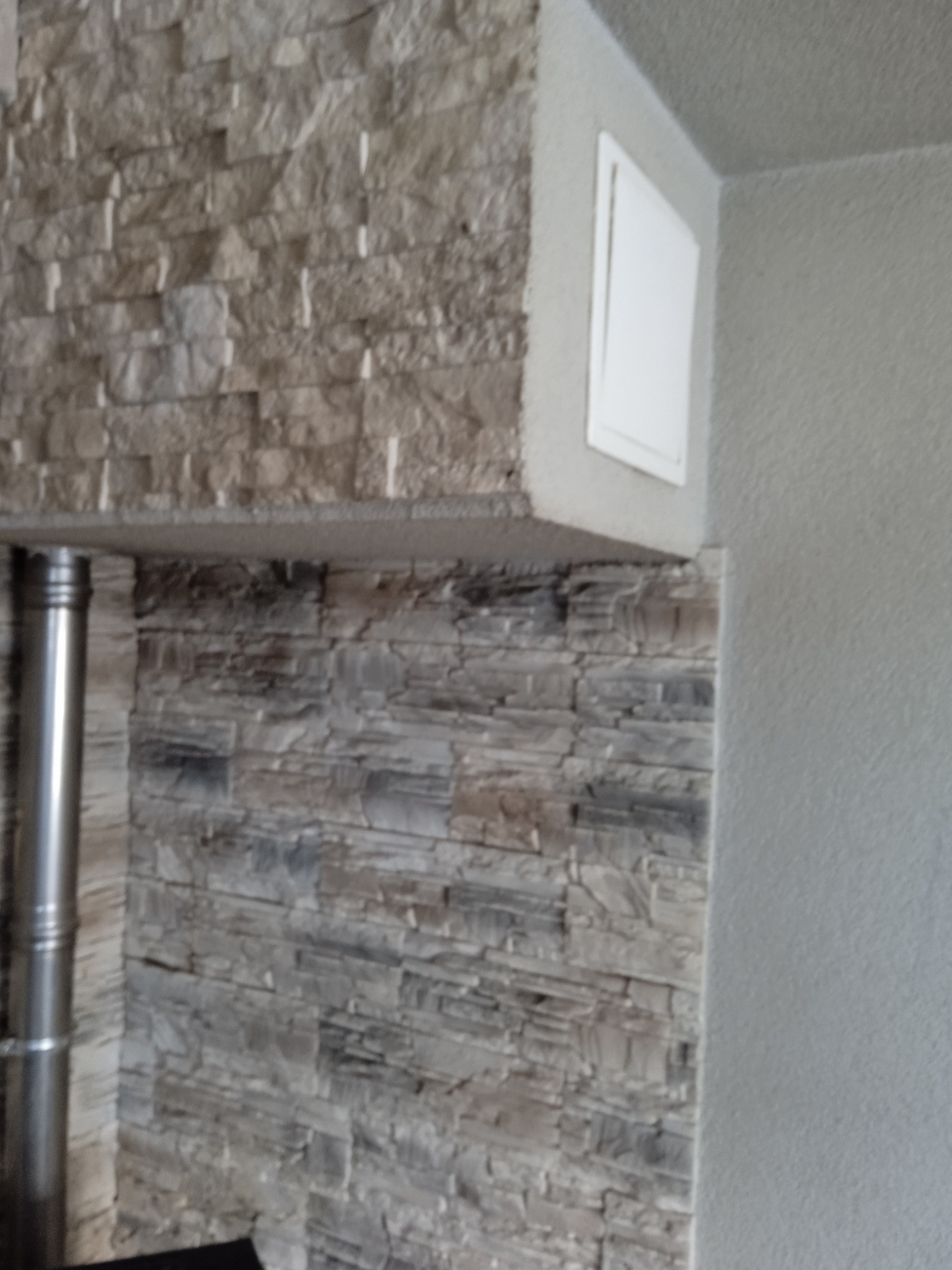 Ściana wykończona dekoracyjnym kamieniem w odcieniach szarości, z widocznym kominem i białą klapą rewizyjną w rogu pomieszczenia.