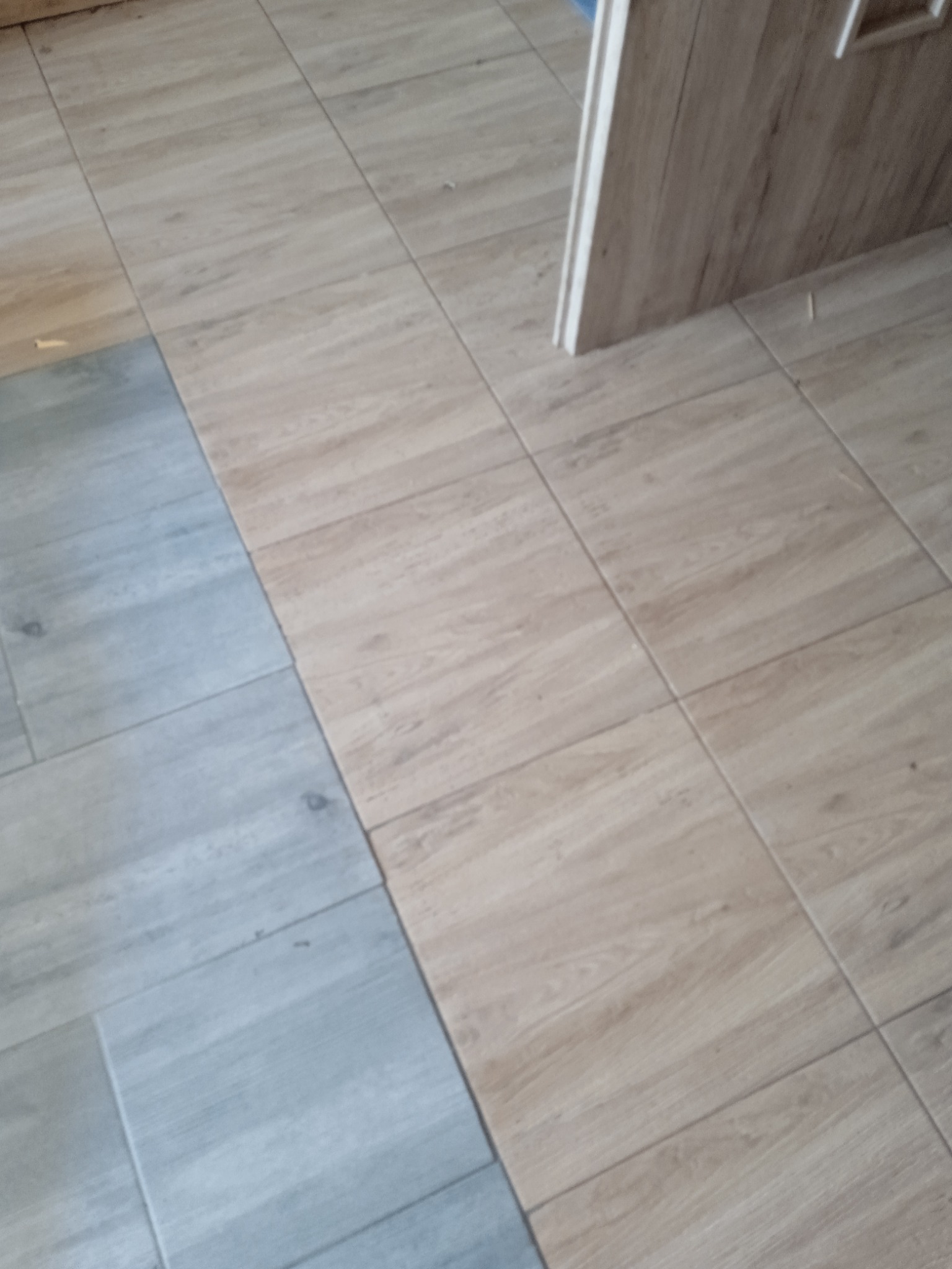 Podłoga wyłożona dwoma rodzajami płytek imitujących drewno: jasnobrązowe i szare. Widoczny fragment ościeżnicy drzwi.