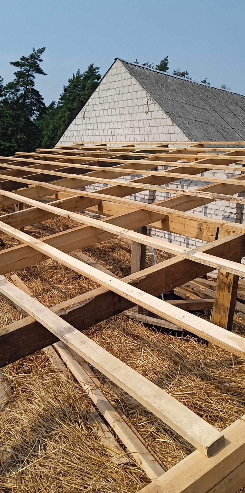 Konstrukcja drewniana dachu w trakcie montażu, widoczne belki i deski na tle muru z białej cegły i fragmentu dachu pokrytego szarą dachówką.