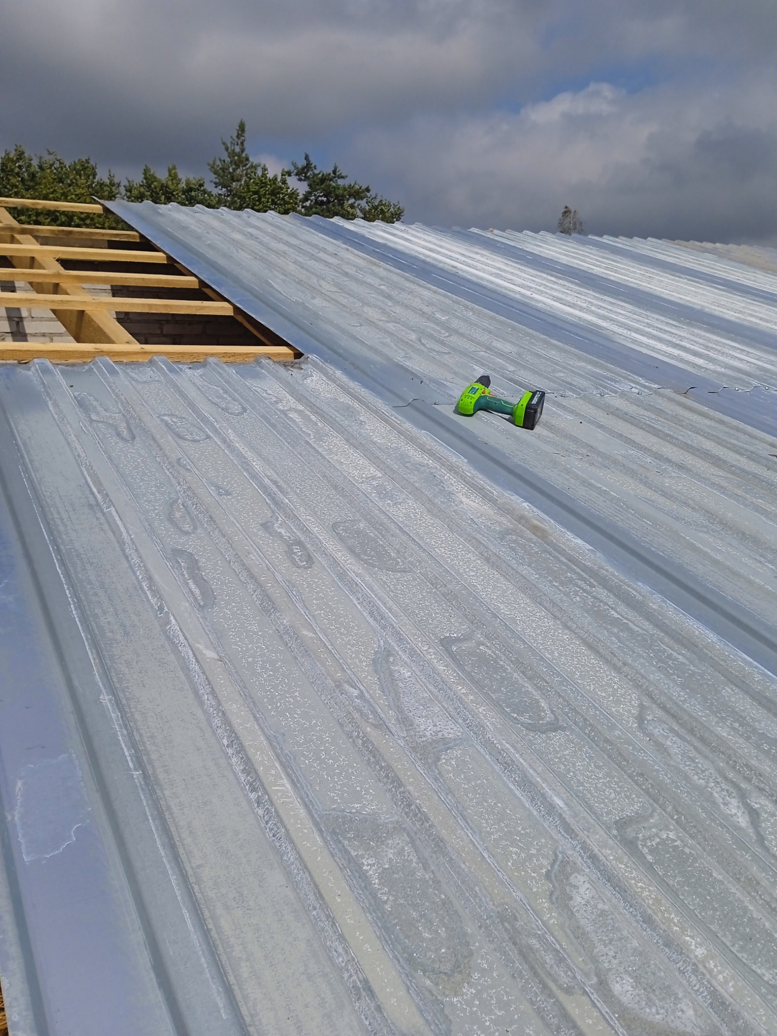 Montaż blachy trapezowej na dachu domu, widoczne krokwie i wkrętarka akumulatorowa leżąca na blasze.