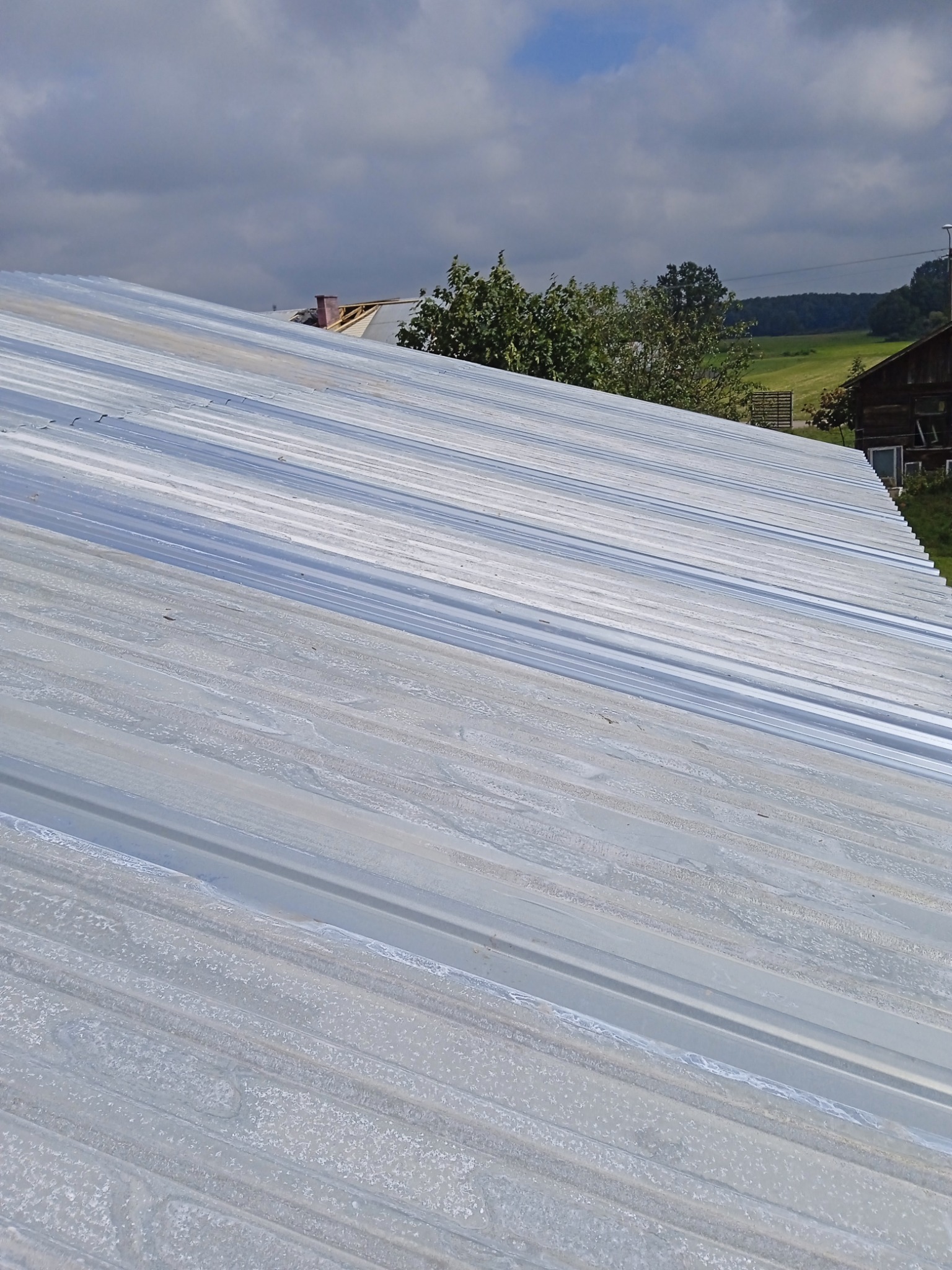 Metalowa powierzchnia dachu z widocznymi łączeniami, lekko zabrudzona, na tle zieleni i błękitnego nieba z chmurami.