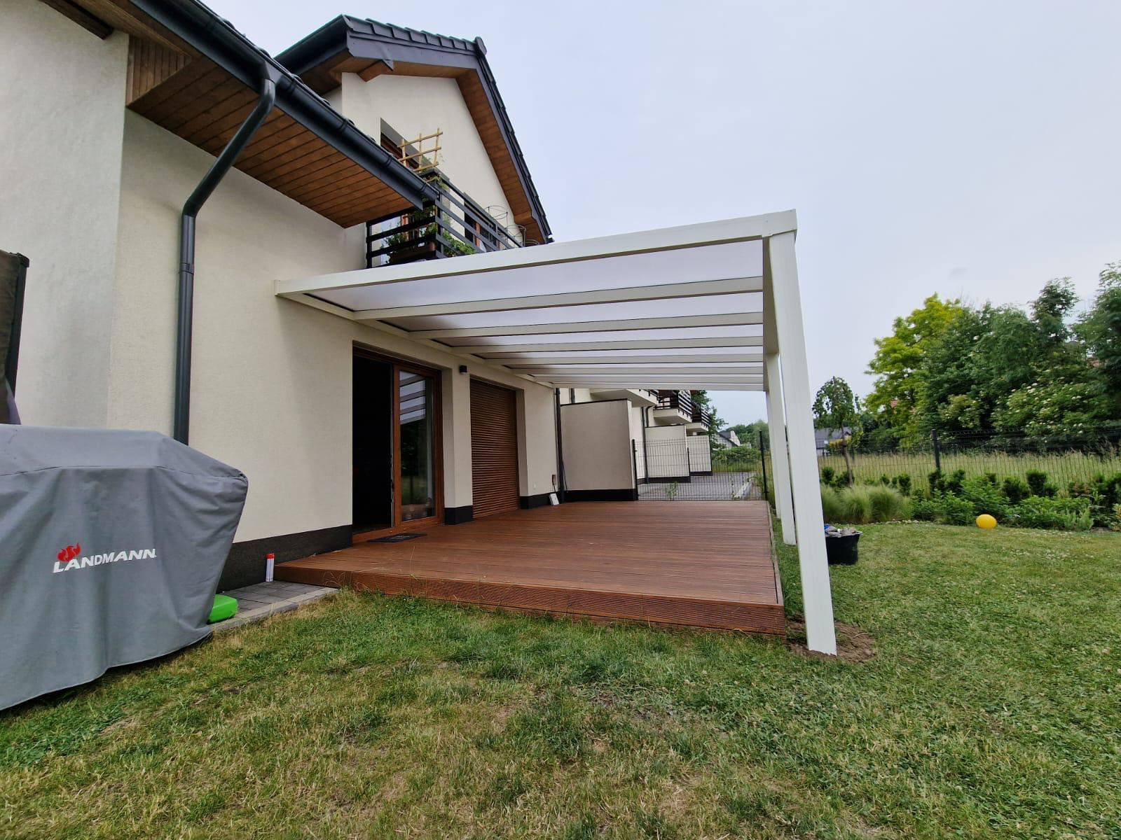 Nowoczesna, biała konstrukcja z zadaszeniem nad drewnianym tarasem przylegającym do domu z brązowymi oknami i roletami, widok z ogrodu.