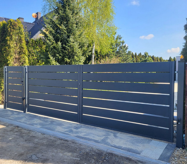 Nowoczesna, przesuwna brama metalowa w kolorze grafitowym z poziomymi szczelinami, zamontowana na posesji z kostki brukowej, w tle zieleń drzew i dom.