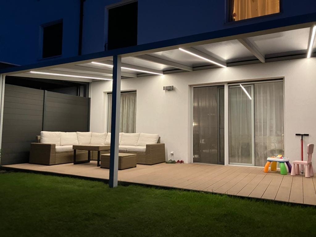 Zadaszony taras z oświetleniem LED, meblami ogrodowymi z rattanu i widokiem na trawnik w nocy.