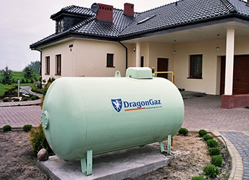 Naziemny zbiornik na gaz płynny marki DragonGaz, umieszczony na betonowej podstawie przed domem jednorodzinnym z jasną elewacją i ciemnym dachem.