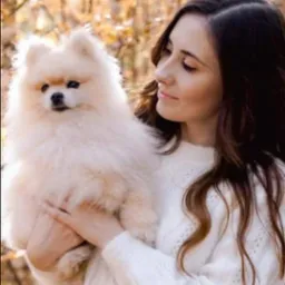 Kobieta w białym swetrze trzymająca na rękach białego szpica miniaturowego na tle jesiennych liści.