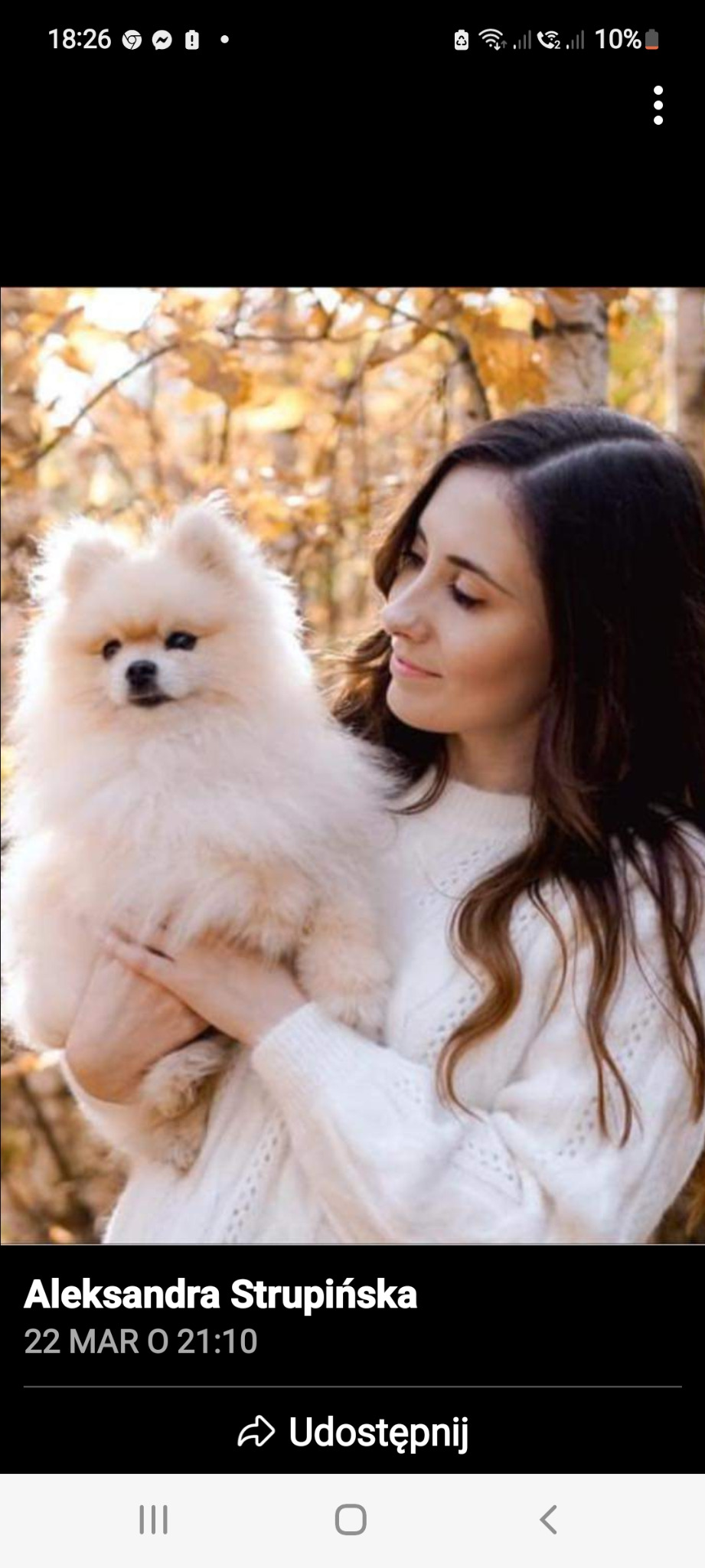 Kobieta w białym swetrze trzymająca na rękach białego szpica miniaturowego na tle jesiennych liści.