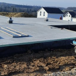 APBud - Betonowa płyta fundamentowa z izolacją pionową, widoczna zagęszczarka wibroprądu i koparka w tle, teren budowy w słoneczny dzień.