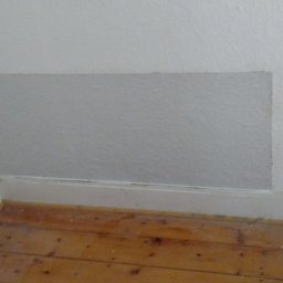 APBud - Fragment ściany po łataniu gładzią, widoczny poziomica i tuba z silikonem w czerwonym wiaderku na podłodze z desek