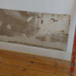 APBud - Fragment ściany z uszkodzoną warstwą farby i widocznymi śladami napraw, poziomica pionowo przy ścianie, drewniana podłoga i listwa przypodłogowa.