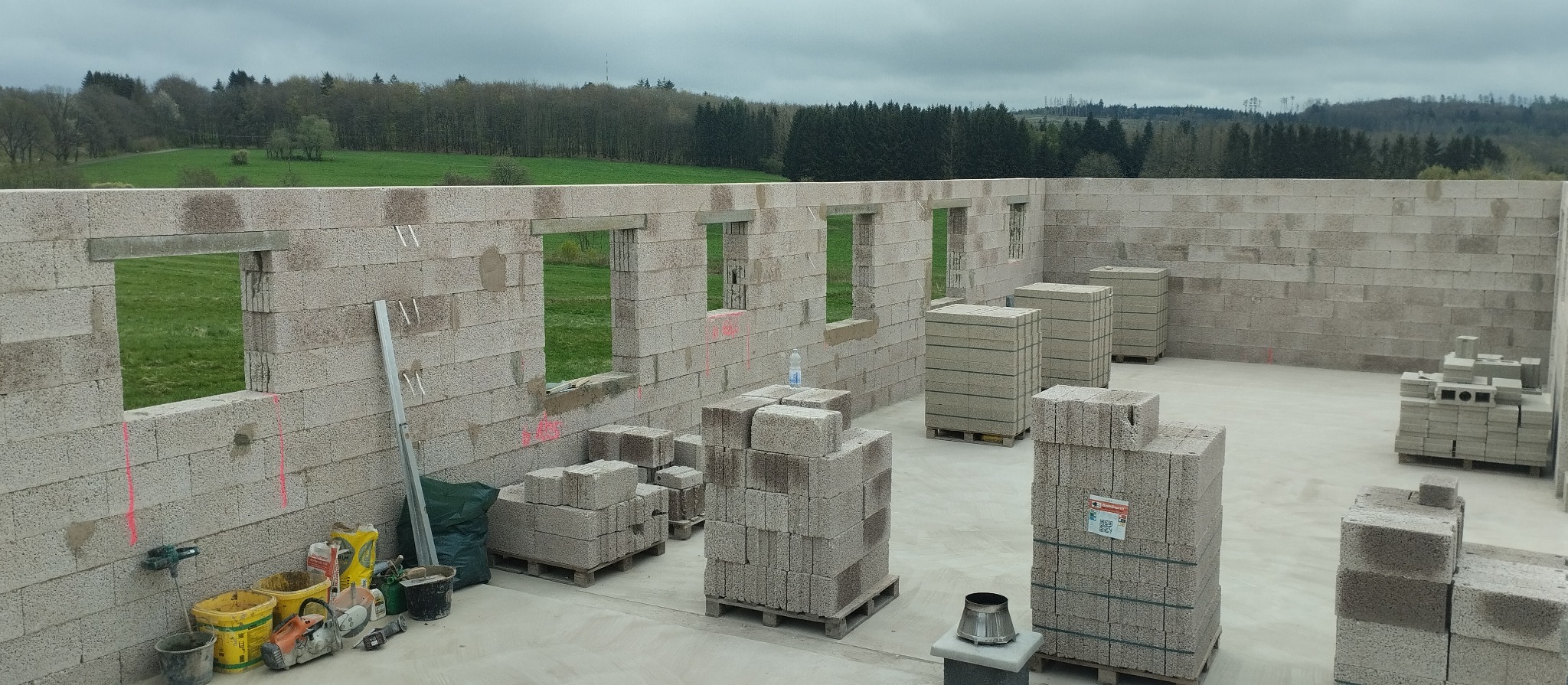 Wznoszenie ścian budynku z bloczków, widoczne otwory okienne, palety z bloczkami, narzędzia budowlane i butelka wody na tle zielonego krajobrazu.