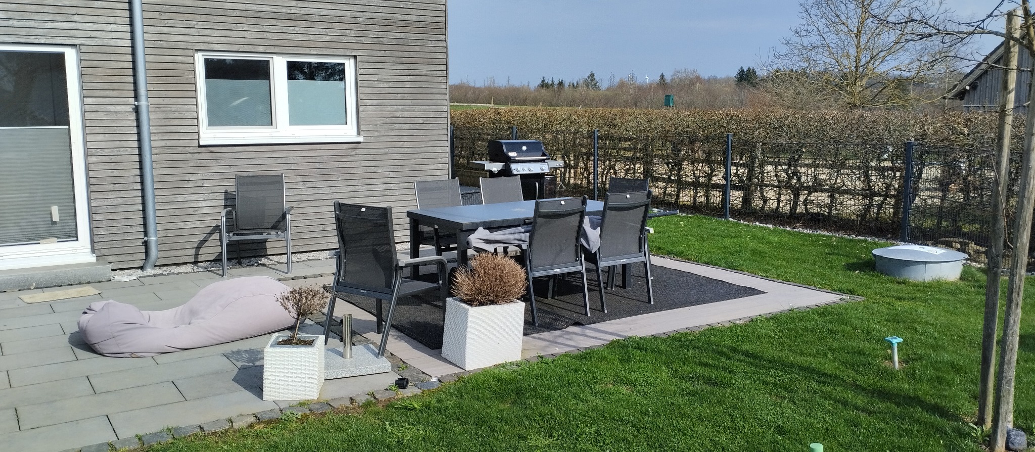 Nowoczesny taras z szarymi panelami elewacyjnymi, szarym zestawem mebli ogrodowych, grillem i pufą w słoneczny dzień.