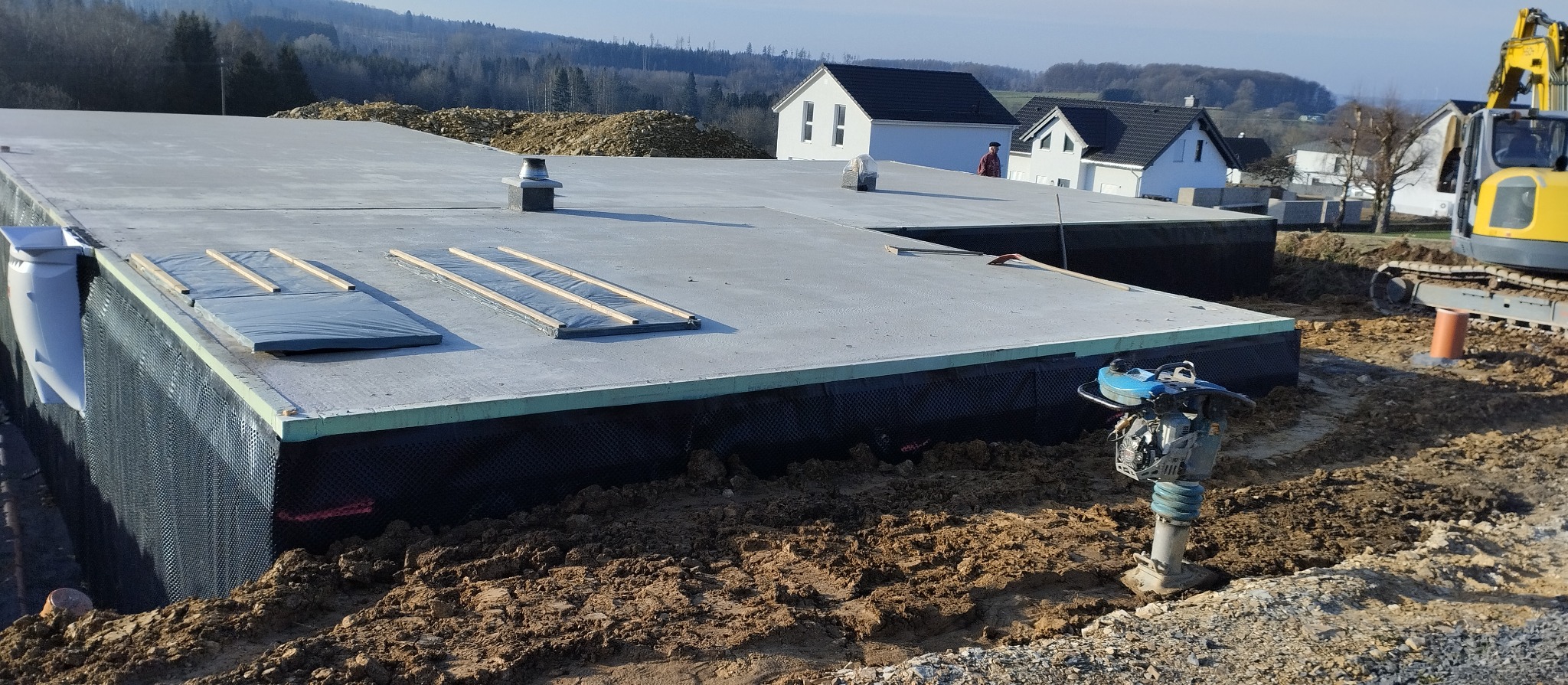 Betonowa płyta fundamentowa z izolacją pionową, widoczna zagęszczarka wibroprądu i koparka w tle, teren budowy w słoneczny dzień.