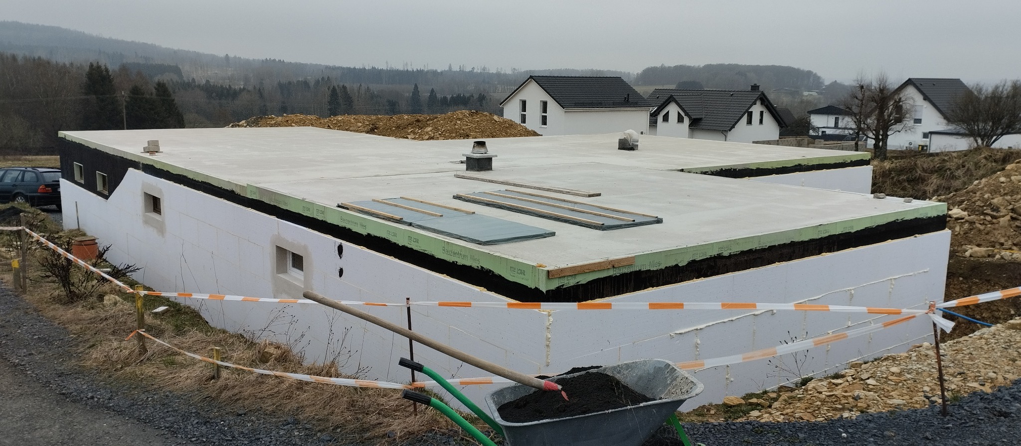 Fundament budynku z białego styropianu, zabezpieczony czarną folią i zielonymi panelami, widoczne okienka, w tle domy i las, w pierwszym planie taczka z ziemią i łopata.