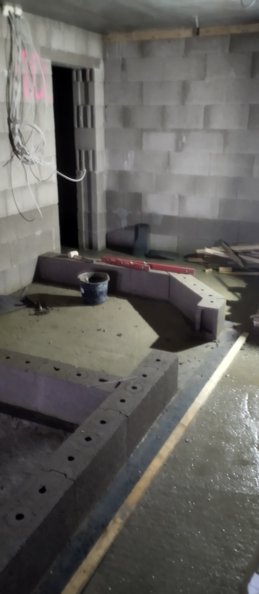 Surowy stan budowy: Wnętrze z szarymi ścianami z bloczków, widoczne przewody elektryczne, przygotowywana wylewka pod podłogę z obudową z pustaków szalunkowych i poziomica w tle.