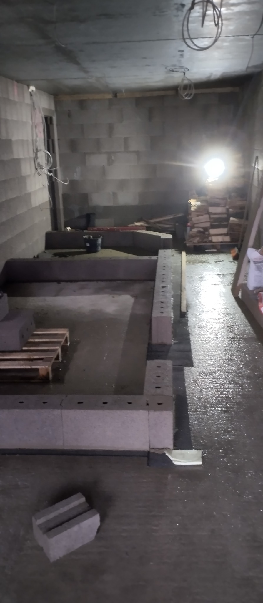 Surowy stan budowy: Ściany z szarego betonu komórkowego w trakcie wznoszenia, widoczne palety, wiadro i prowizoryczne oświetlenie w pomieszczeniu.