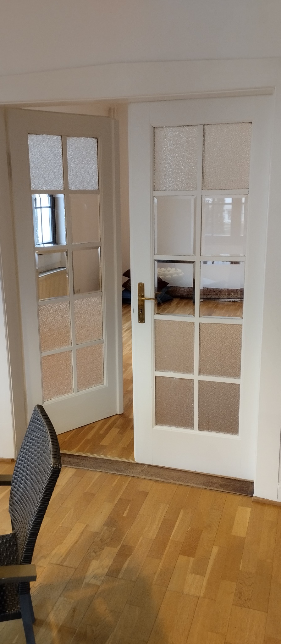 Białe, przeszklone drzwi wewnętrzne z drewnianą podłogą w mieszkaniu, widok na pokój z meblami przez otwarte skrzydło.