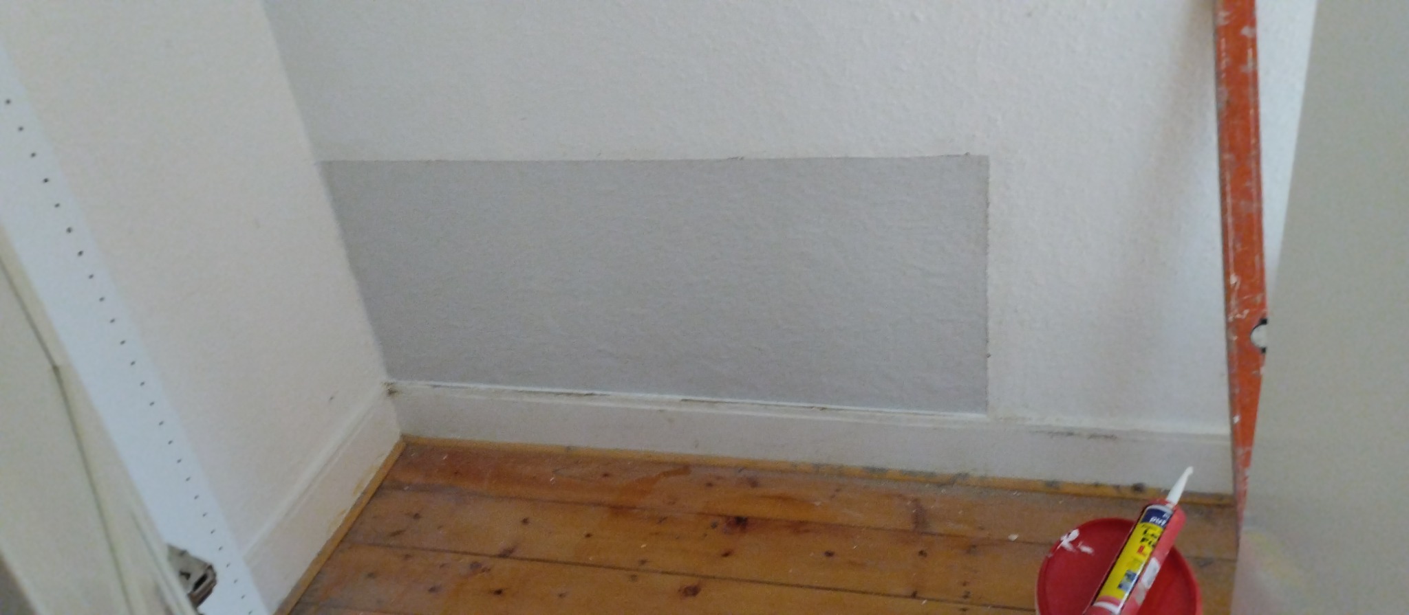 Fragment ściany po łataniu gładzią, widoczny poziomica i tuba z silikonem w czerwonym wiaderku na podłodze z desek