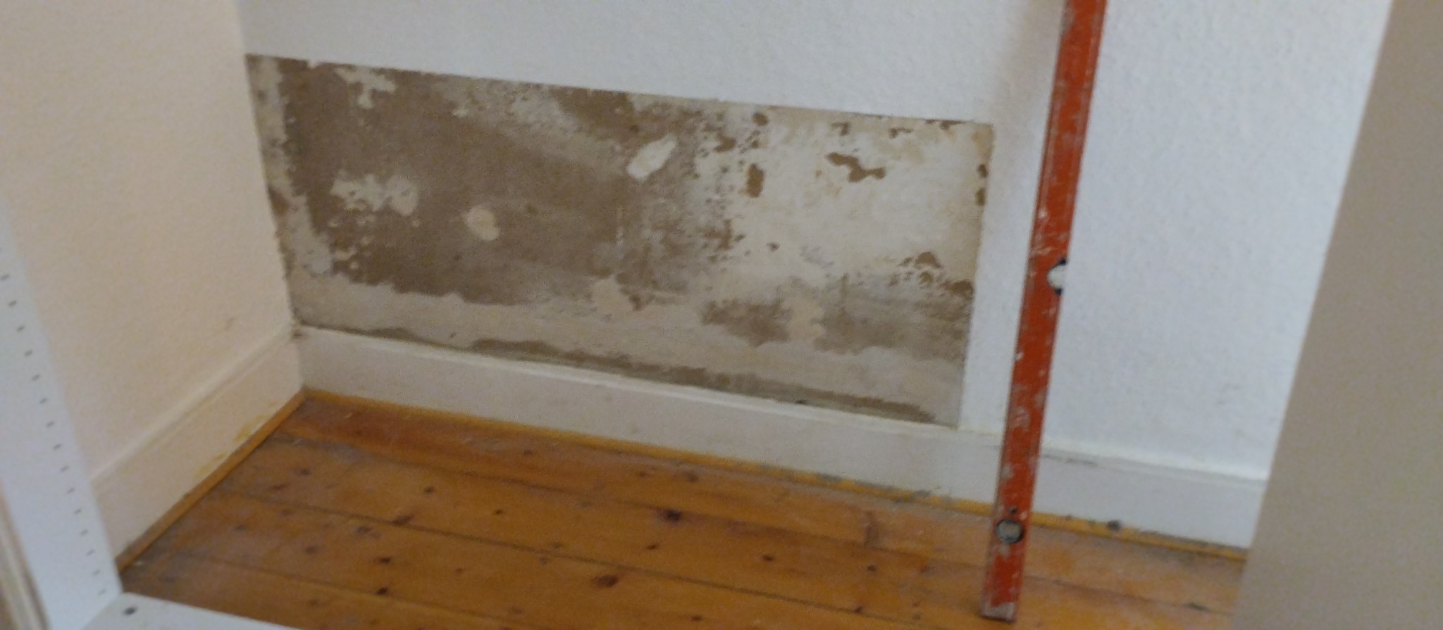 Fragment ściany z uszkodzoną warstwą farby i widocznymi śladami napraw, poziomica pionowo przy ścianie, drewniana podłoga i listwa przypodłogowa.