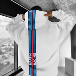 Biała bluza z kapturem marki Sparco z pionowym pasem Martini Racing na plecach, widok z tyłu na tle miejskiego krajobrazu za oknem.