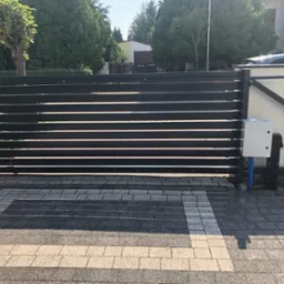 Nowoczesna, pozioma brama przesuwna z automatyką, zamontowana na posesji z kostki brukowej, widoczna puszka sterownicza i niebieski peszel.