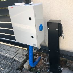 IntelAmper - Biała skrzynka sterownicza z niebieskim gniazdem i niebieską rurą karbowaną, zamontowana przy bramie wjazdowej, obok czarnego słupka z zamkiem i kluczem.