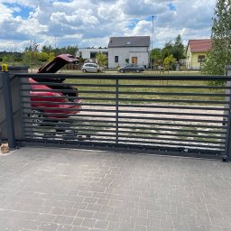 IntelAmper - Szara, przesuwna brama z automatycznym napędem, widoczny silnik i lampa ostrzegawcza, z otwartym bagażnikiem czerwonego samochodu w tle, na podjeździe z kostki brukowej.