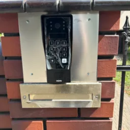 Zamontowany wideodomofon EURA z klawiaturą numeryczną, umieszczony na ceglanym słupku ogrodzenia z skrzynką na listy. Widoczne śruby mocujące i fragment metalowej bramy.