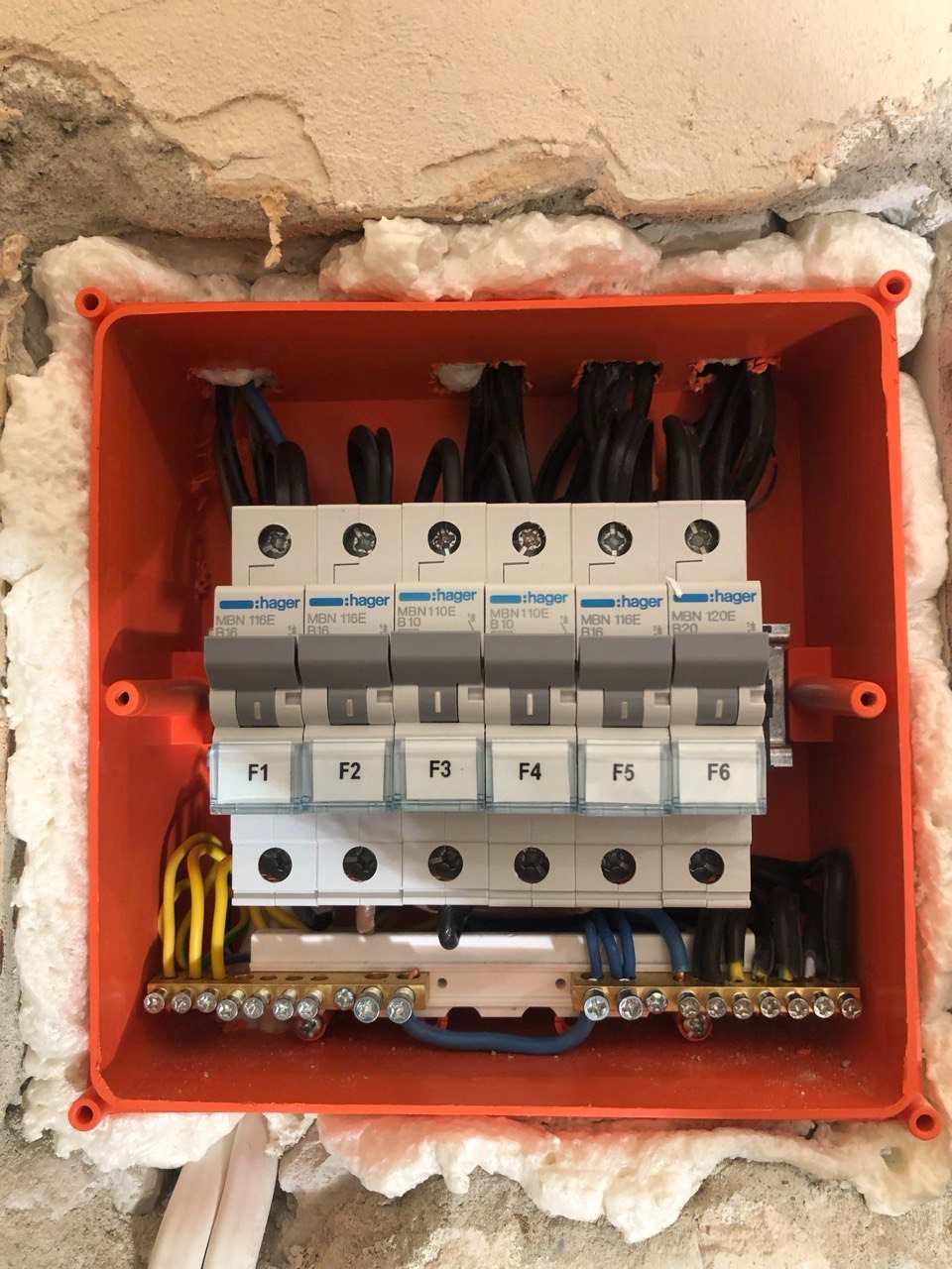 Pomarańczowa skrzynka rozdzielcza Hager z zainstalowanymi bezpiecznikami i przewodami, zamontowana w ścianie z widoczną pianką montażową wokół obudowy.