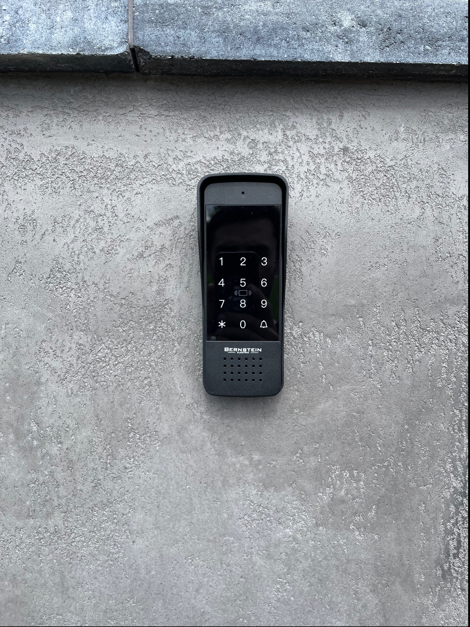 Zamontowany na szarej, chropowatej ścianie, czarny panel domofonu z klawiaturą numeryczną i logo 'BERNSTEIN ELECTRIC'.