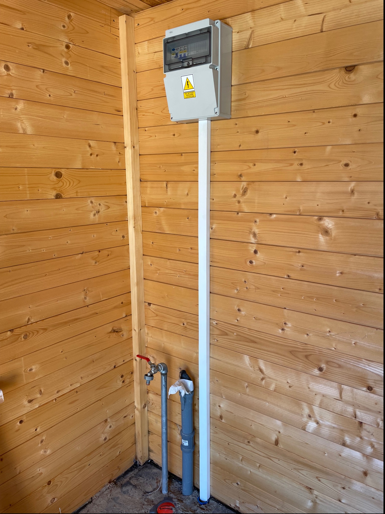 Szara skrzynka elektryczna z żółtą naklejką ostrzegawczą zamontowana na drewnianej ścianie z boazerii, biała listwa maskująca przewody, obok rura wodna z kranem i rura kanalizacyjna.