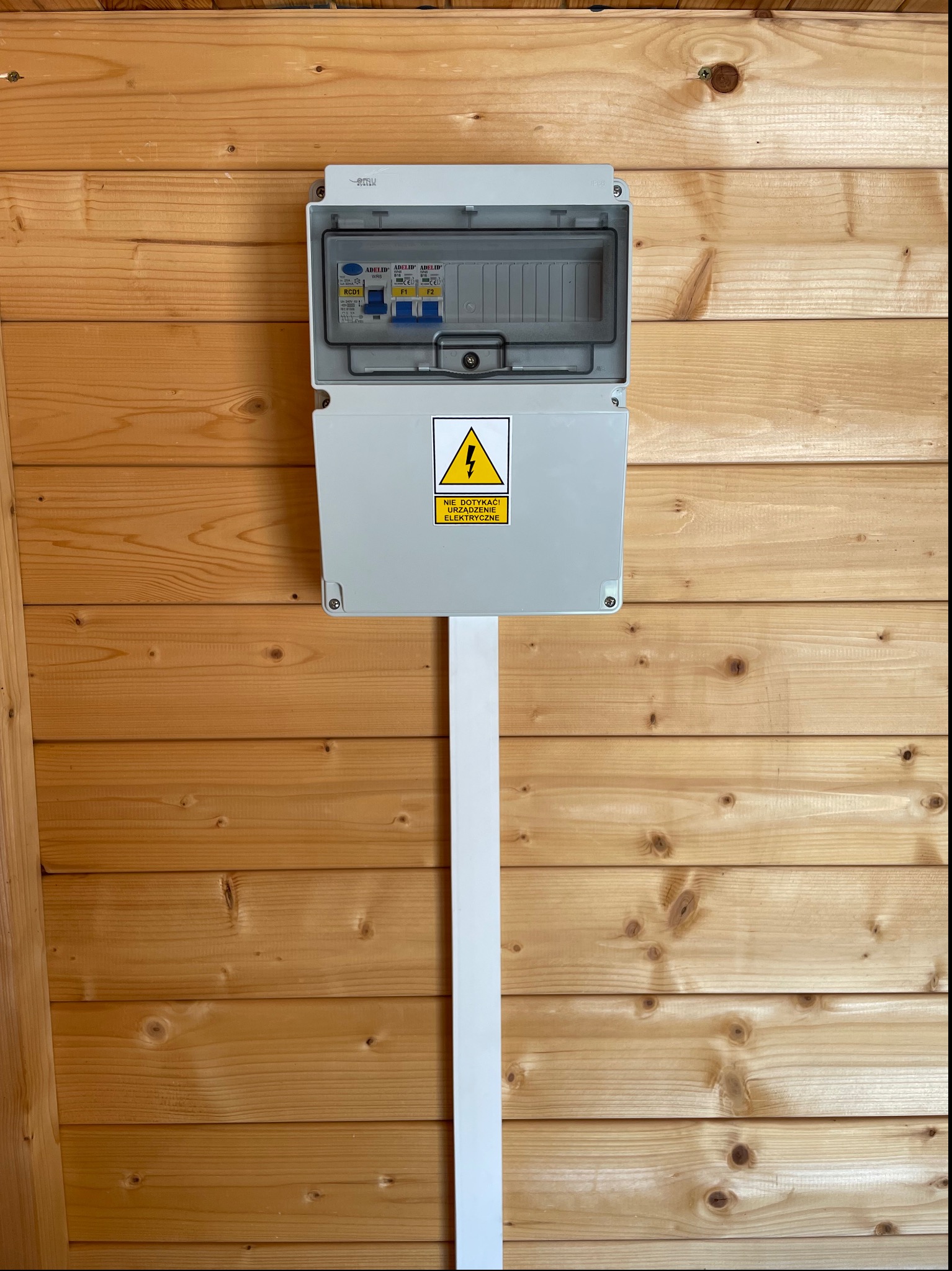 Otwarta skrzynka elektryczna z widocznymi bezpiecznikami, zamontowana na ścianie z boazerii, z żółtą naklejką ostrzegawczą o napięciu.