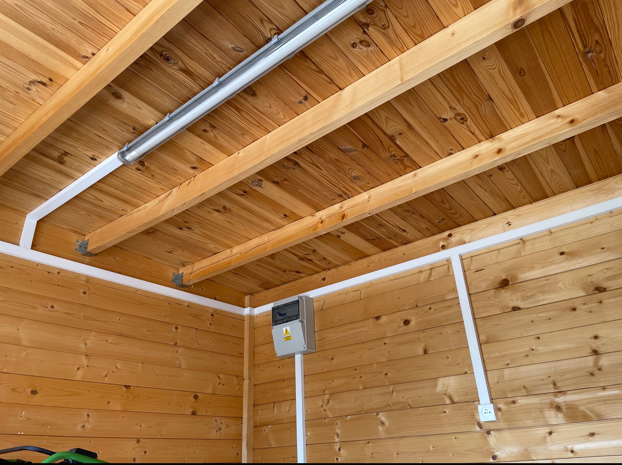 Wnętrze drewnianego pomieszczenia z widoczną skrzynką elektryczną z symbolem ostrzegawczym, zamontowaną na ścianie, oraz zainstalowaną lampą jarzeniową pod drewnianym sufitem.