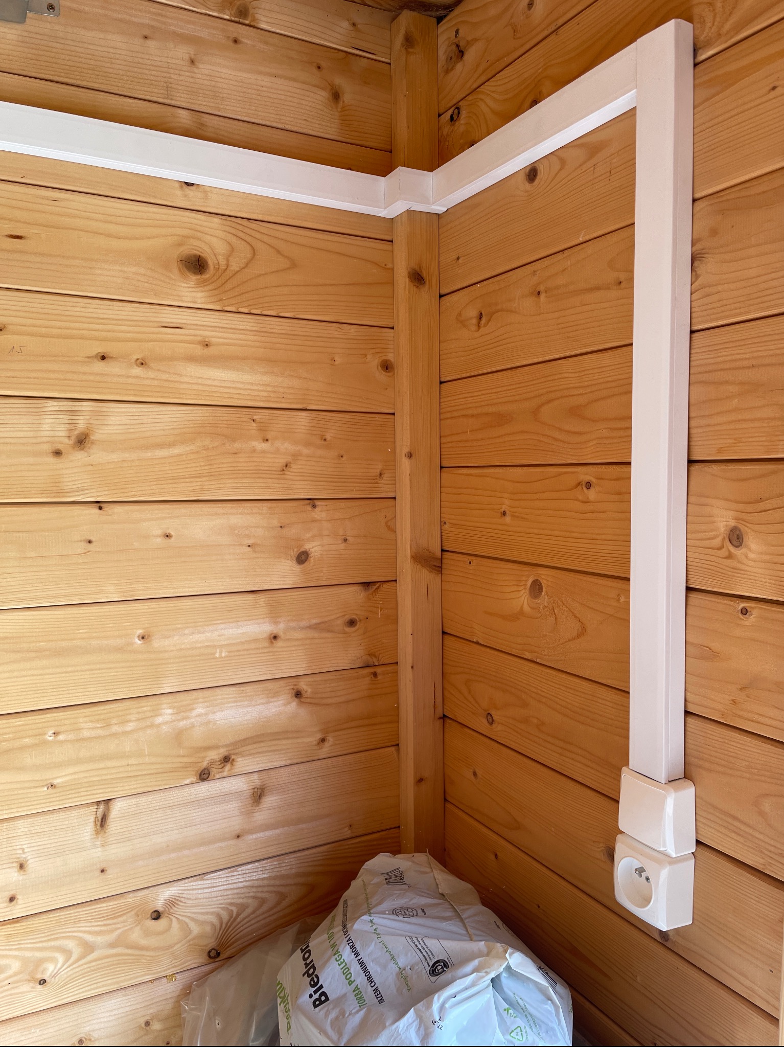 Kąt pomieszczenia wyłożonego boazerią z jasnego drewna z poprowadzoną białą listwą przypodłogową i pionową rurą osłonową instalacji elektrycznej zakończoną podwójnym gniazdkiem, u dołu widoczna...