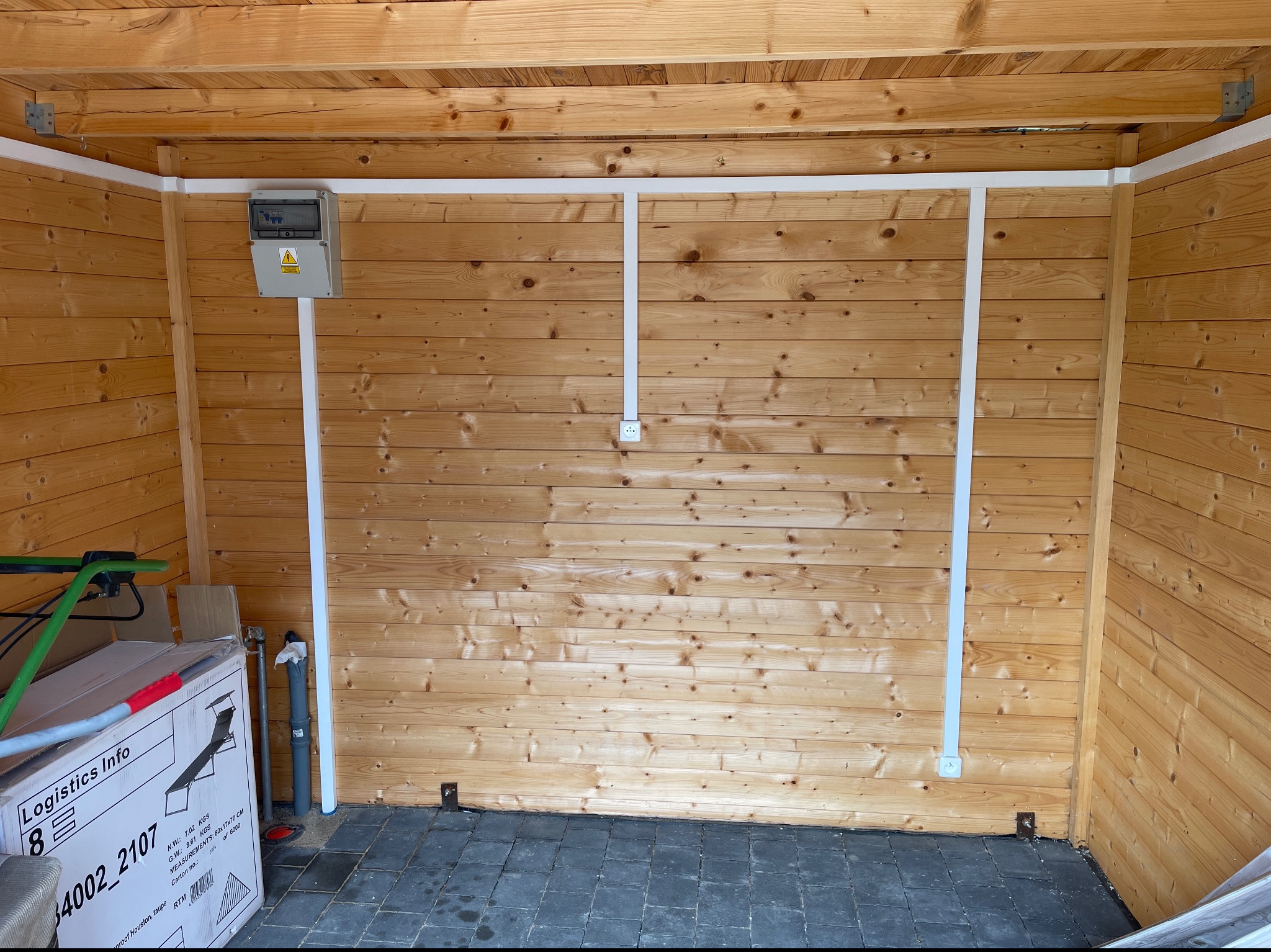 Wnętrze drewnianej altany w Poznaniu z zamontowaną skrzynką elektryczną, białymi listwami maskującymi przewody i gniazdkiem. Na podłodze szara kostka brukowa, w rogu karton z napisem Logistics Info.