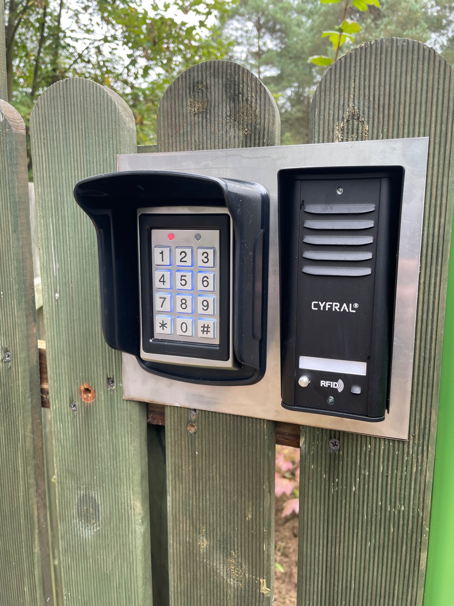 Zewnętrzny panel domofonowy Cyfral z klawiaturą numeryczną i czytnikiem RFID zamontowany na drewnianej bramie.