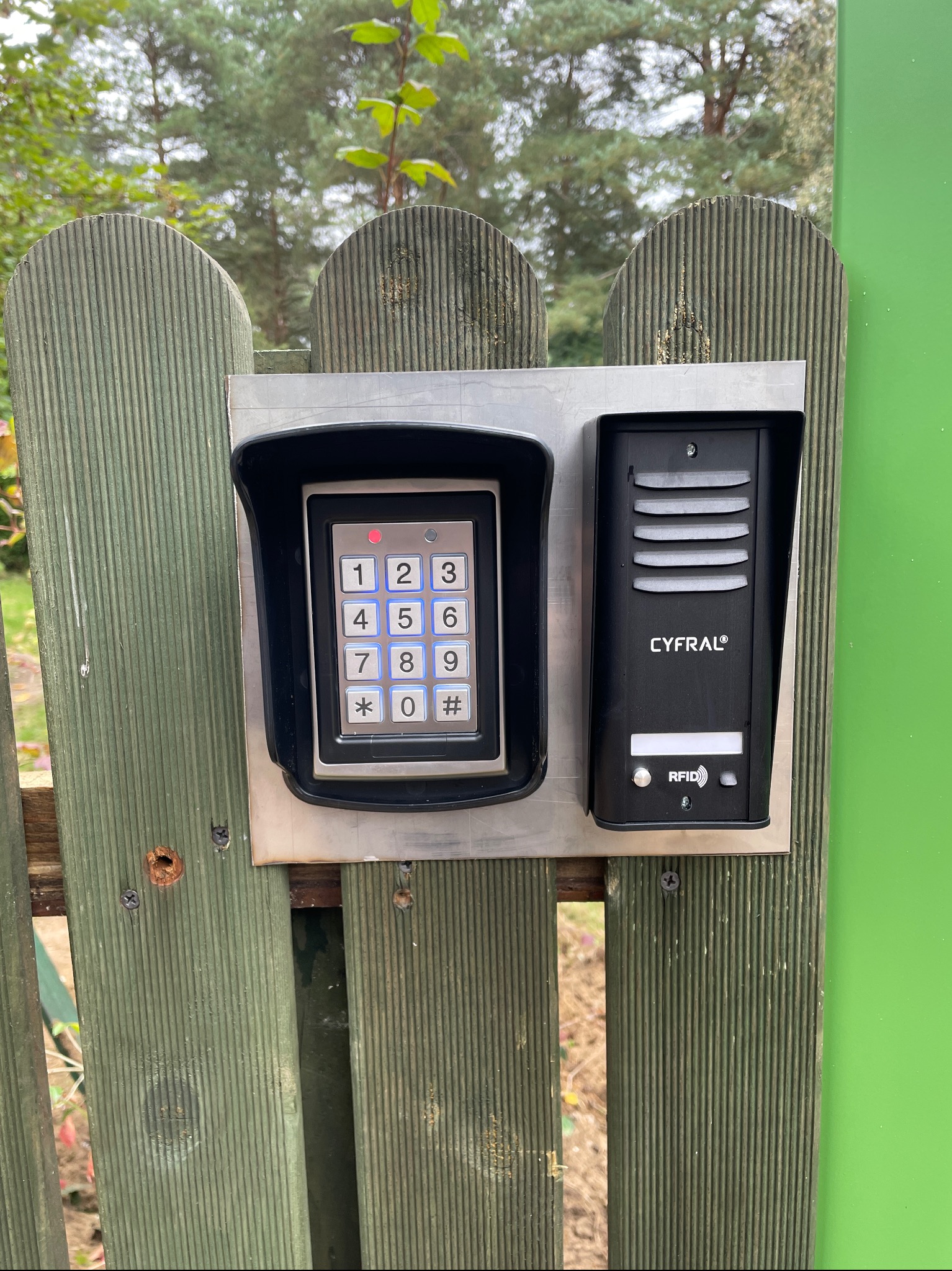 Zewnętrzny panel domofonowy z klawiaturą numeryczną i czytnikiem RFID marki Cyfral, zamontowany na drewnianej furtce pomalowanej na zielono, z widocznymi śladami użytkowania i otoczeniem zieleni.