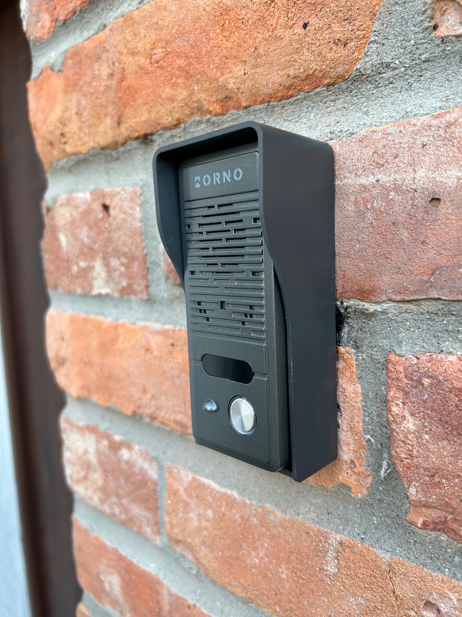 Czarny, nowoczesny domofon marki ZORNO zamontowany na ceglanej ścianie zewnętrznej budynku.