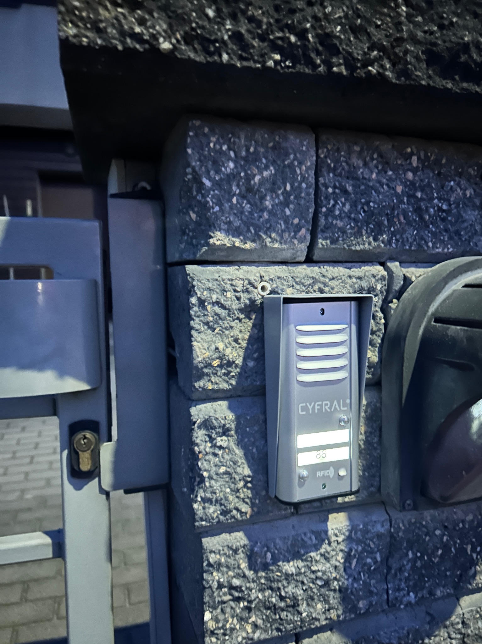 Szary domofon Cyfral zamontowany na murze z betonowych bloczków, obok skrzynki na listy i furtki.