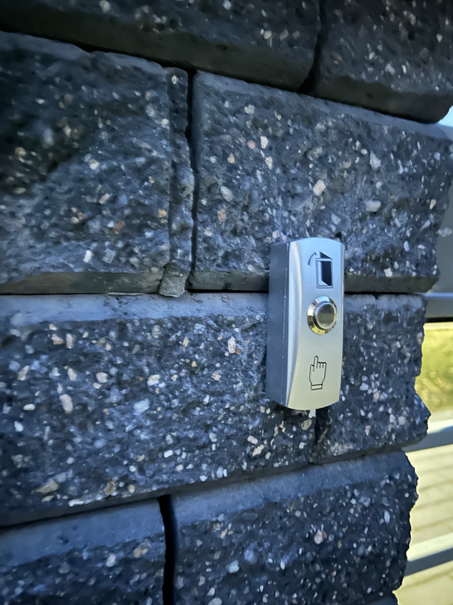 Srebrny panel domofonu z przyciskiem i symbolem dłoni wskazującej na tle szarego, kamiennego muru.