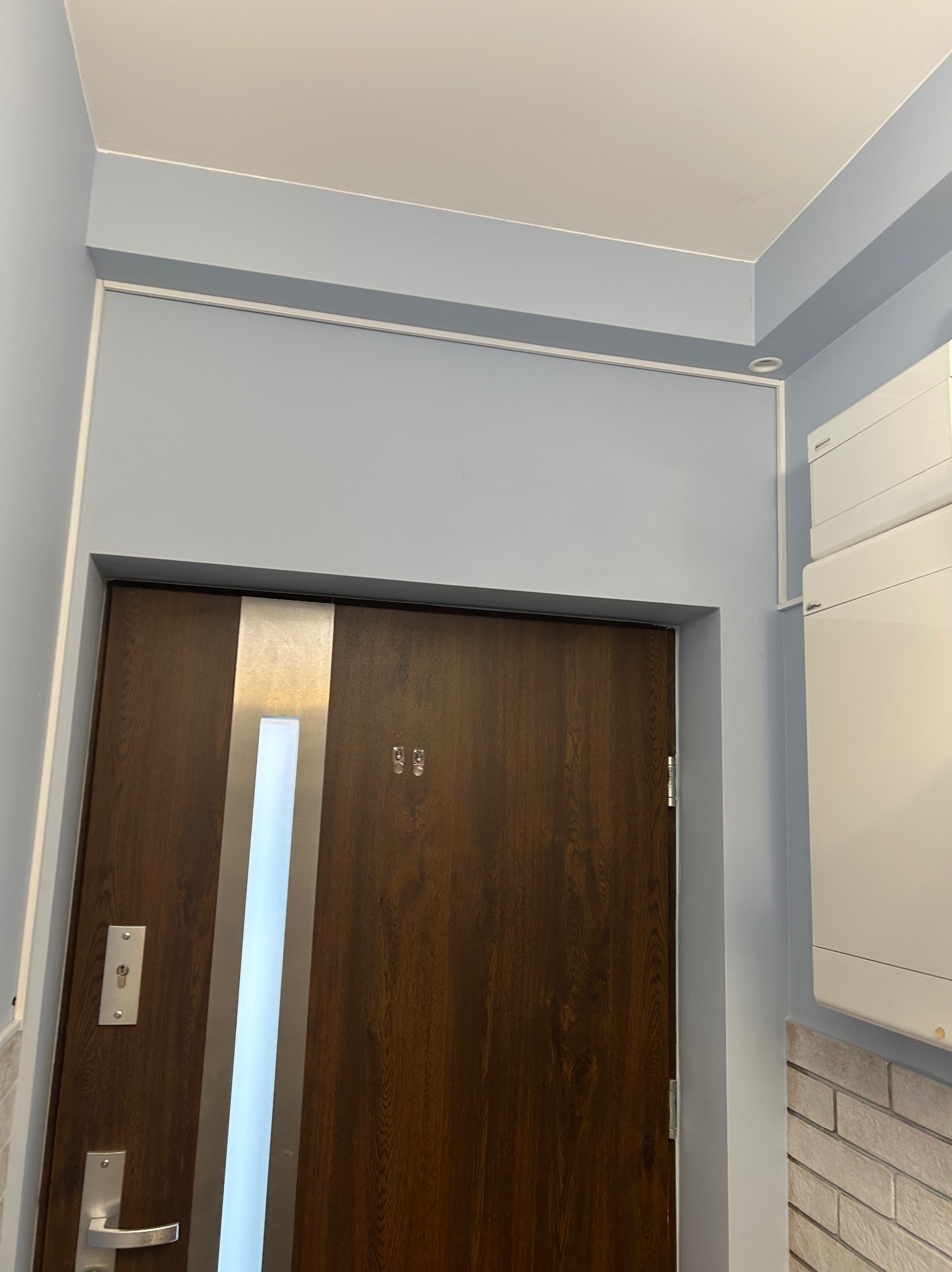 Białe listwy nadtynkowo prowadzące kable elektryczne wzdłuż sufitu i ściany, obok brązowych drzwi z metalową wstawką i białym panelem.