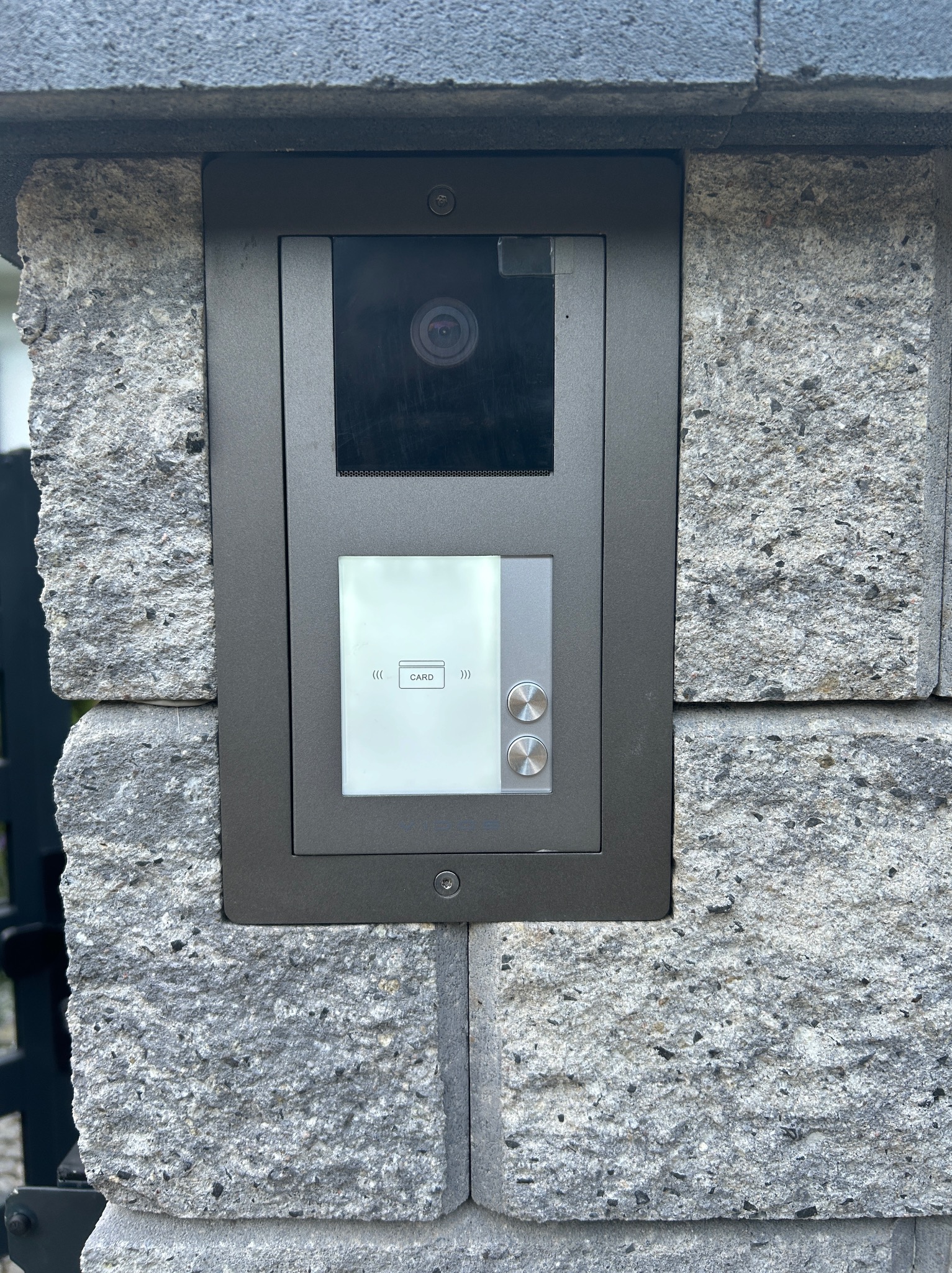 Nowoczesny wideodomofon z czytnikiem kart zbliżeniowych zamontowany na kamiennym słupku ogrodzenia, Poznań. Szara obudowa, kamera i podświetlany ekran.